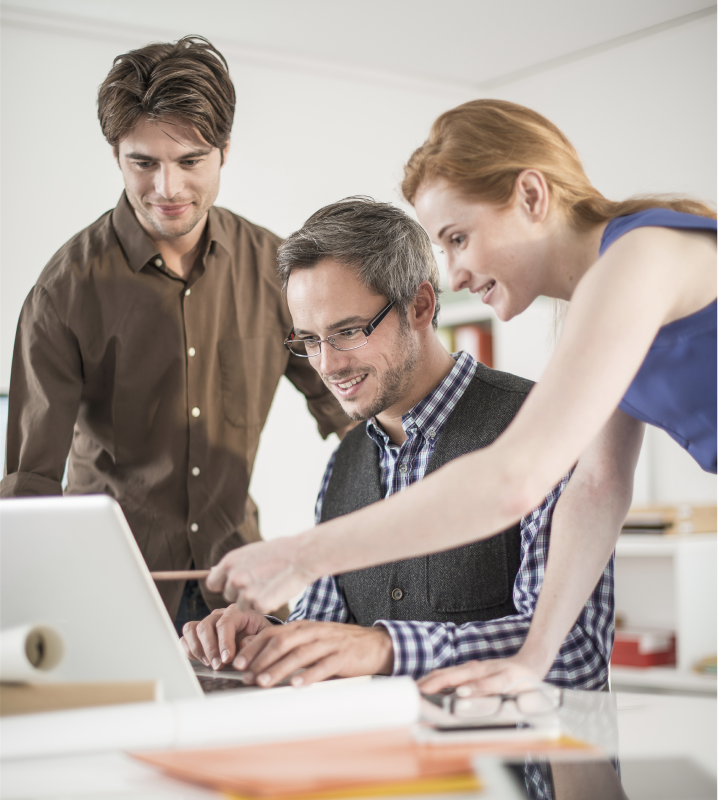 Three colleagues collaborating at a desk, looking at a laptop screen with engaged expressions.